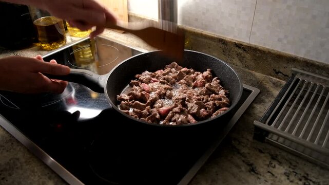 Close-up of raw beef cubes being fried in a pan with a wooden spatula, cooking process in a home kitchen.