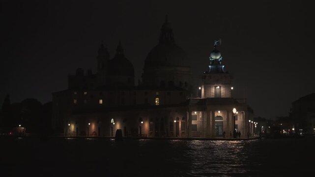 Punta della Dogana and Santa Maria della Salute Basilica at night in Venice. View from a moving boat on the water. Illuminated historic architecture with reflections on the lagoon surface.