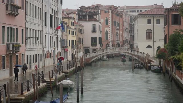 People walk with umbrellas along a Venice canal on a rainy day. A gondola passes under a historic arched bridge. Authentic Italian cityscape with wet pavement and colorful old buildings.