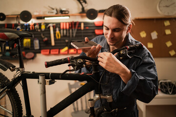 Male bike mechanic with mobile phone talk speakerphone record voice message on smartphone recording voicemail working in bike shop or garage