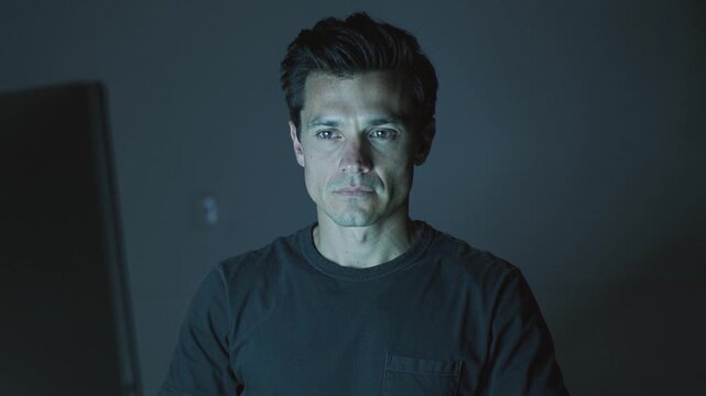 Focused man sits at desk staring at computer monitor in dark room. Illuminated by screen light, he represents overtime work, cybersecurity, gaming or programming late at night.