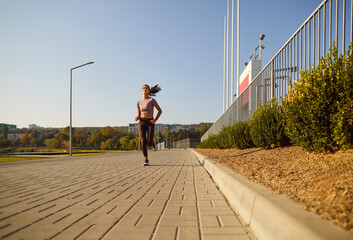Obraz premium Woman runner jogging outdoors in park. She runs on a paved walkway in warm daylight, symbolizing cardio training, endurance fitness, and wellness goals. Energy, focus, and wellness concept.