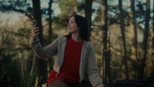  a young woman hiking in a forest as she video calls with her smartphone. With a bright smile and a backpack, she captures the moment amidst the natural scenery, reflecting the joy 