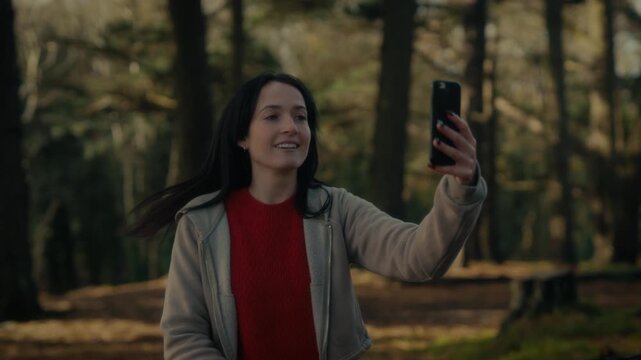 a young woman hiking in a forest as she video calls with her smartphone. With a bright smile and a backpack, she captures the moment amidst the natural scenery, reflecting the joy 