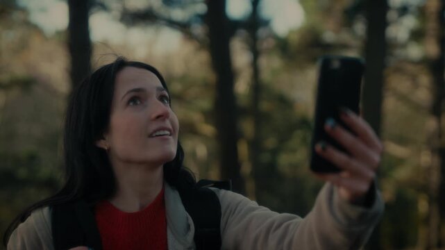  a young woman hiking in a forest as she video calls with her smartphone. With a bright smile and a backpack, she captures the moment amidst the natural scenery, reflecting the joy 