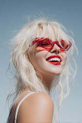 Cheerful Woman on Summer Vacation Pointing at Camera &ndash; Red Sunglasses and Lipstick on Blue Background