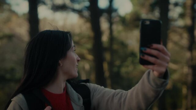  a young woman hiking in a forest as she video calls with her smartphone. With a bright smile and a backpack, she captures the moment amidst the natural scenery, reflecting the joy 