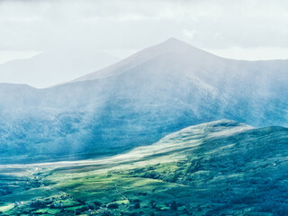 The rolling green landscape of County Kerry, Ireland. The hills and valleys are illuminated by sunlight, while ominous clouds create a dramatic atmosphere above the mountains.