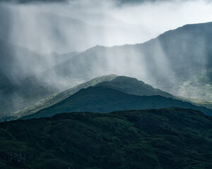 Dark green mountains are shrouded in mist and rain. Streaks of sunlight break through the clouds, illuminating sections of the mountain landscape.