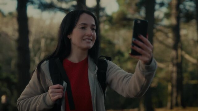  a young woman hiking in a forest as she video calls with her smartphone. With a bright smile and a backpack, she captures the moment amidst the natural scenery, reflecting the joy 