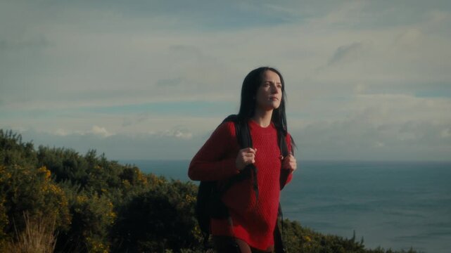  a young woman standing at a scenic viewpoint, gazing out over a tranquil body of water and distant hills. With her backpack slung over her shoulders.
