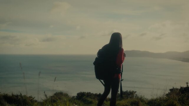  a young woman standing at a scenic viewpoint, gazing out over a tranquil body of water and distant hills. With her backpack slung over her shoulders.
