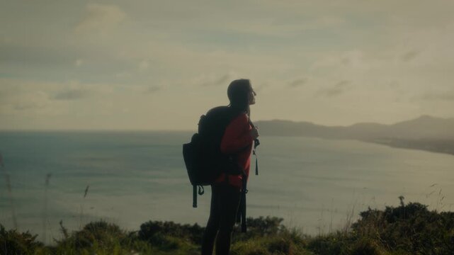  a young woman standing at a scenic viewpoint, gazing out over a tranquil body of water and distant hills. With her backpack slung over her shoulders.