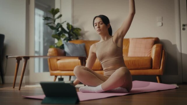 woman practising yoga in a cosy living room setting. She is in a lunging pose on a yoga mat, with a tablet in front of her displaying a workout or meditation session. 