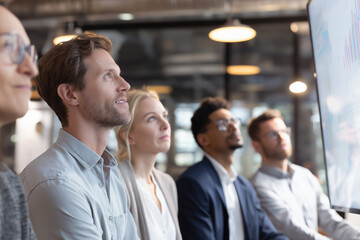 A diverse group of business professionals managers attentively listens as a confident speaker presents data on a digital display during a productive business meeting.