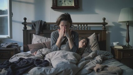 Young woman sitting on bed rubbing eyes while partner sleeps. She looks exhausted and stressed suffering from insomnia or waking up early. Concept for sleep disorders and health care. - Powered by Adobe