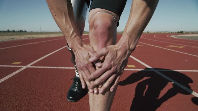 Close up disabled athlete with prosthetic leg holding knee on track. Man feeling pain or warming up before training outdoors. Concept sport disability resilience.