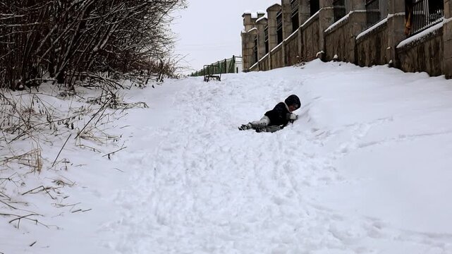 A joyful little boy slides down a small snowy hill during a winter walk. The child enjoys active outdoor play, demonstrating sincere emotions of happiness, winter fun, and an atmosphere of carefree ch