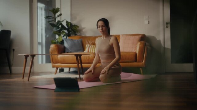 woman practising yoga in a cosy living room setting. She is in a lunging pose on a yoga mat, with a tablet in front of her displaying a workout or meditation session. 
