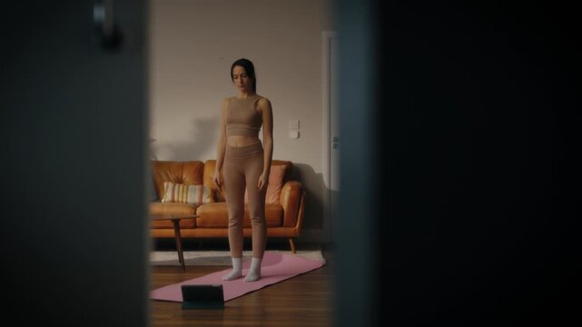 woman practising yoga in a cosy living room setting. She is in a lunging pose on a yoga mat, with a tablet in front of her displaying a workout or meditation session. 