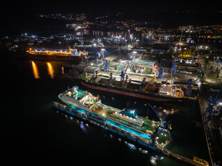 Night aerial photo of an industrial port with shipyard facilities, cranes, docks and illuminated vessels. Urban harbor infrastructure glowing with city lights along the waterfront