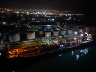Aerial night photo of an industrial port with an oil tanker moored at the dock, illuminated storage tanks and refinery infrastructure along the waterfront, city lights reflecting on the water