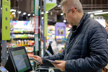 A man in a black puffer jacket uses his smartphone to pay at a self-checkout station in a supermarket. He focuses on the screen, completing his transaction with modern digital convenience.