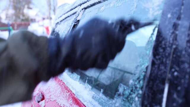 A gloved hand scrapes ice off a car window in a snowy environment. The scene captures the essence of winter weather and vehicle maintenance.