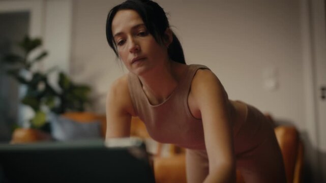 woman practising yoga in a cosy living room setting. She is in a lunging pose on a yoga mat, with a tablet in front of her displaying a workout or meditation session. 