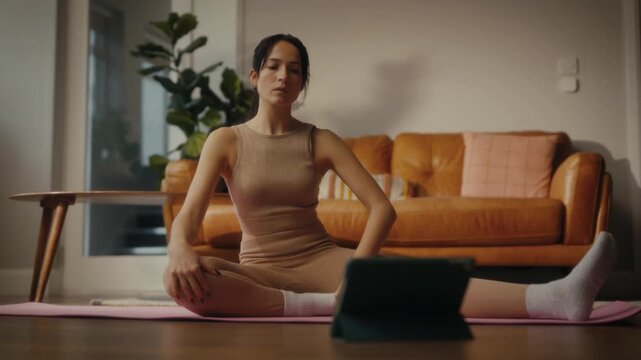 woman practising yoga in a cosy living room setting. She is in a lunging pose on a yoga mat, with a tablet in front of her displaying a workout or meditation session. 