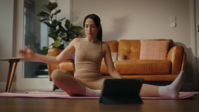 woman practising yoga in a cosy living room setting. She is in a lunging pose on a yoga mat, with a tablet in front of her displaying a workout or meditation session. 