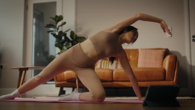 woman practising yoga in a cosy living room setting. She is in a lunging pose on a yoga mat, with a tablet in front of her displaying a workout or meditation session. 