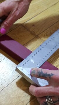 A close-up of a tattooed hand with pink nails using a metal L-square ruler and a pencil to draw a precise straight line on a red wooden beam during a DIY project.