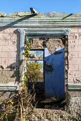 Weathered wall with doorway and crumbling plaster, revealing vegetation and blue sky through an open ruin in bright daylight.
