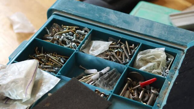 High angle shot of an open blue plastic hardware organizer box divided into sections, containing various types of metal screws, wall plugs, and fasteners for home repair.