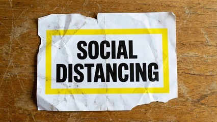 Social Distancing Notice: A close-up shot of a ripped piece of paper that gives a very important safety warning against infection. It has a yellow border and placed on the wooden background.