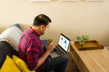 Adult man working from home, eating sandwich and analyzing data on laptop while sitting on couch, combining remote work and lunch