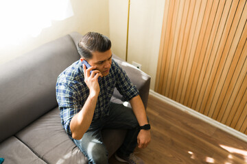 High angle view of a man sitting on couch and talking on smartphone, looking concerned during an...