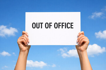 Two raised hands hold a white paper sign with Out of Office text with clear blue sky.