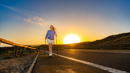 Beautiful middle-aged woman practicing nordic walking on seaside road on summer sunrise. Front view	