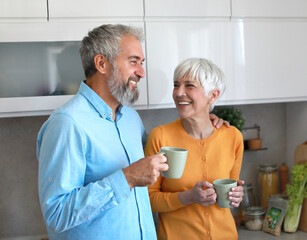 A man and a woman are smiling and holding cups of coffee in a kitchen. Scene is happy and friendly