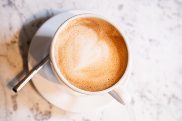 birds eye view of cappuccino in a white coffee mug on a saucer, with a spoon, on a white marble table