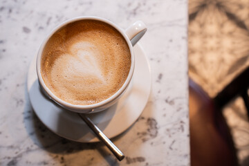birds eye view of cappuccino in a white coffee mug on a saucer, with a spoon, on a white marble table