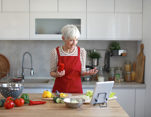 Portrait of happy senior mid aged mature woman prepering meal with fresh vegatebles and following internet instructions for a recipe on a tablet computer in kitchen