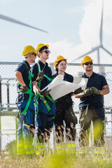 Technician team wearing safety harnesses and hardhats holding blueprints at a wind farm site....