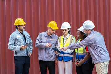 Diverse group of professional logistics engineers and workers standing in front of a red shipping container, having a team meeting and discussing operations using a digital tablet and walkie-talkie.