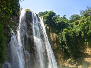 A majestic waterfall cascades down a lush rocky cliff, viewed from behind a rustic wooden railing under a bright blue sky with fluffy white clouds, capturing the serene beauty of nature. © Iruzzayne