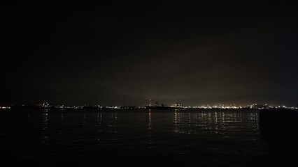 A wide night shot captures illuminated ships anchored in a dark harbor, with city lights shimmering across the calm water under a moody cloudy sky.