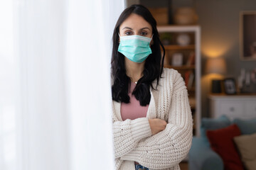 Woman wearing a protective face mask standing indoors near a window and looking at the camera. Lifestyle healthcare image focused on safety, prevention, and health awareness.
