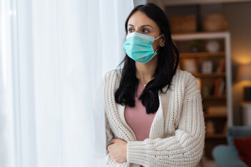 Woman wearing a protective face mask standing near a window at home and looking outside. Lifestyle healthcare image focused on safety, prevention, and health awareness.
